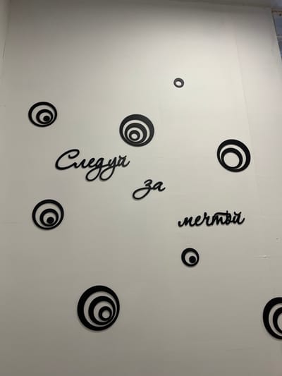 This is a photograph of a white wall in an indoor environment, possibly a room in Калодзішчанскі сельскі Савет, Belarus. The wall is decorated with black, concentric circle decals and stylized black script. The script reads "Следуй за мечтой" which translates to "Follow your dream". There are nine concentric circle decals of varying sizes distributed across the wall. Three of the decals are larger, with multiple rings, and are positioned on the left and bottom portions of the image. Six smaller decals, with fewer rings, are scattered throughout the upper and right portions of the wall. The script is positioned in the central area of the wall, with the words "Следуй" and "за" in the upper left and middle respectively, and "мечтой" on the right. The lighting appears to be from an overhead source, casting subtle shadows from the decals.