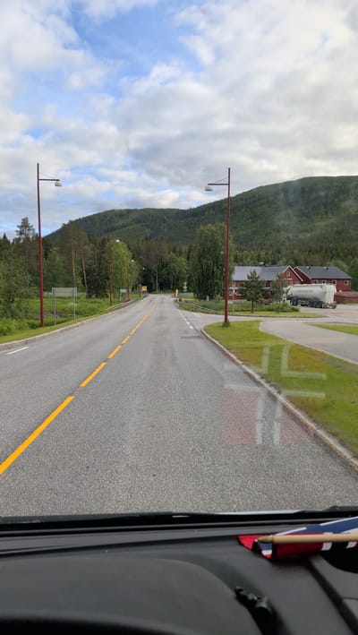 The image depicts a road in Namsskogan, Norway. The road is paved and has a double yellow line dividing the lanes. It curves to the left and is bordered by green grass and trees. In the background, there are hills covered in dense green forest. On the right side of the road, there are red buildings with white trim and a white truck with a tank trailer. Several red streetlights are visible along the road. The sky is partly cloudy with a mix of blue sky and white clouds. The photo appears to be taken from inside a vehicle, with the dashboard and a small flag visible in the foreground.