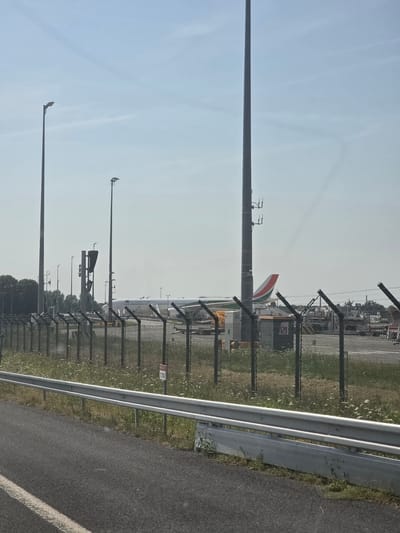 The image shows an outdoor scene at an airport, likely in Roissy-en-France, France, given the context of large commercial aircraft. A large white passenger airplane, with "Air Côte d'Ivoire" visible on its fuselage, is parked on the tarmac. The airplane has a distinctive tail fin with green, white, and orange stripes. Several tall light poles with multiple fixtures are visible throughout the scene, along with a traffic signal with a dark housing. A chain-link fence with barbed wire runs along the foreground, separating the viewer's perspective from the airport grounds. Beyond the fence, there is an area with grass and small white flowers. A metal guardrail is positioned along the edge of an asphalt road in the immediate foreground. In the background, there are trees and other airport support vehicles and equipment. The sky is clear and blue with a few wispy clouds.
