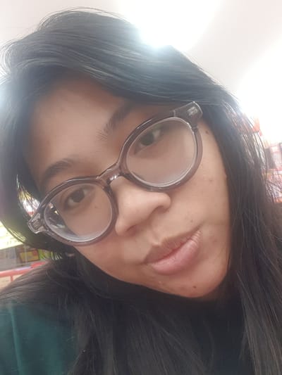 A close-up, downward-angled selfie of a young woman with dark hair and glasses. She is wearing a dark green top. The background appears to be an indoor setting, possibly a store, with shelves and products visible. The lighting is bright, with some glare on the glasses. The woman's face is turned slightly to the left, and her mouth is slightly open. The image is focused on her face and upper body.