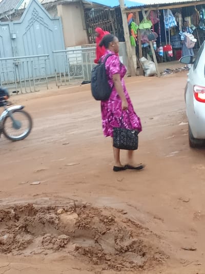 The image depicts an outdoor scene on a dirt road in Kagini, Nigeria. A woman wearing a vibrant pink patterned dress and carrying a black backpack walks across the road, holding a patterned black handbag in her right hand. She is wearing dark flat shoes. Behind her, a motorcycle is partially visible, with its front wheel in motion. In the background, there is a market stall with various items for sale, including bags and clothing, and a light-colored car is parked on the right side of the frame, with its taillight illuminated. A grey metal gate and fencing are also visible to the left of the woman. The ground is uneven and appears to be a mixture of dirt and mud.