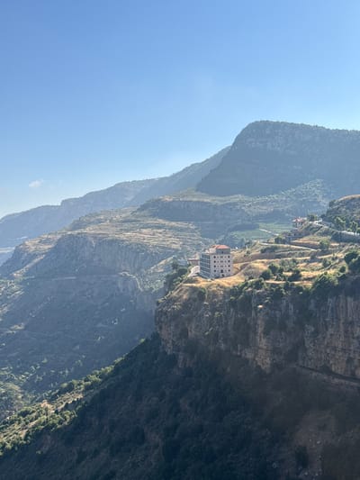 The image shows a scenic view of the mountainous landscape surrounding Jezzine, Lebanon. In the foreground, a dramatic cliff face covered in dark green vegetation drops sharply. Perched on the edge of this cliff is a multi-story building with a red tiled roof, constructed from light-colored stone. The building appears to be modern but blends with the traditional architecture of the region.
Further into the distance, the terrain softens into terraced hillsides, some appearing cultivated with rows of crops or vineyards, while others are covered in dry, golden grass and scattered trees. More buildings, smaller and with red roofs, are visible nestled into these slopes, suggesting a village or hamlet. A winding road is visible traversing the landscape, connecting the various structures and the different levels of the hills.
The background is dominated by layers of majestic, hazy mountains stretching towards the horizon under a clear, pale blue sky. The sunlight casts shadows, highlighting the contours and ruggedness of the terrain. The overall impression is one of natural beauty, a harmonious blend of human habitation and the imposing mountain environment.