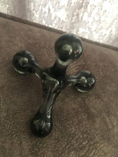 A dark, translucent, cross-shaped object made of glass or resin lies on a brown textured surface, possibly upholstery. The object has a central stem with three rounded protrusions, one at the top, one to the left, and one to the right. The surface it rests on appears to be a couch or chair, with a blurred, light-colored fabric in the background. The lighting casts reflections on the object, highlighting its smooth, polished surface. The location of Путёвка, Russia, is not depicted in this image.