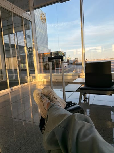 The image displays an interior view of Munich Airport (MUC), located within the Freising district of Germany. The lower half of a person is visible in the foreground, wearing light blue denim pants, white socks, and white platform sneakers with patterned laces. Their feet are elevated and resting on a black hard-shell carry-on suitcase with a silver telescopic handle. To the right, a section of black airport seating with an attached side table is visible. The terminal features large glass windows, through which a golden-hour sky, indicative of sunrise or sunset, is apparent. Outside, a multi-story glass building prominently displays a yellow Lufthansa logo. An airport bus, partially obscured but showing "Omnibusbetrieb" on its front, is parked on the tarmac, surrounded by other ground support equipment and vehicles. The polished, light-colored tiled floor of the terminal reflects the warm natural light entering through the windows.