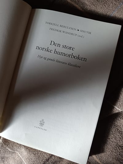 The image shows an open book with a light-colored cover laid on a brown, textured fabric. The book's title, "Den store norske humorboken," is visible in large, dark font. Below the title, in smaller font, it reads "Nye og gamle litterære klassikere." Above the title, the names "TORKJELL BERULFSEN" and "FREDRIK WANDRUP (red.)" are printed, with "SISS VIK" to the right of the first name. A small logo with flowers and the text "CAPPELEN 1829" is located near the bottom of the page. The lighting creates strong shadows across the book and the fabric. There is no indication of the city of Hovden, Norway in the image.