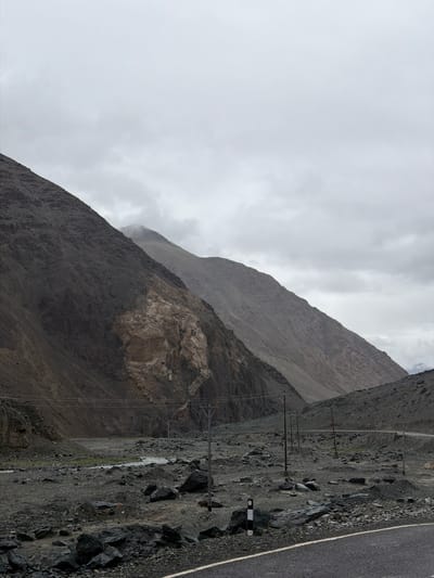 The image depicts a rugged, mountainous landscape with a cloudy sky overhead. The terrain is characterized by steep, rocky slopes and a barren, stony valley floor. A paved road curves through the scene, with a white line marking its edge. Several power poles with wires are visible, indicating human infrastructure in this remote area. In the distance, a small stream or river flows through the valley. The overall atmosphere is somber and desolate. The location is identified as Sumdo, India, a village in the Ladakh region known for its stark, high-altitude environment. No people are present in the image.