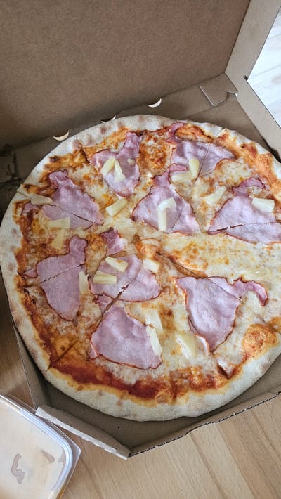 A close-up, overhead shot shows a Hawaiian pizza in a cardboard pizza box, placed on a wooden surface. The pizza is circular with a golden-brown crust, topped with melted cheese, tomato sauce, thinly sliced ham, and chunks of pineapple. The pizza is cut into eight slices. To the left of the pizza box is a clear plastic container with a beige sauce or dip. The pizza box is open, and its cardboard interior is visible. The background is a light-colored wooden floor. The scene appears to be an indoor setting in the city of Slengiai, Lithuania, likely a kitchen or dining area.