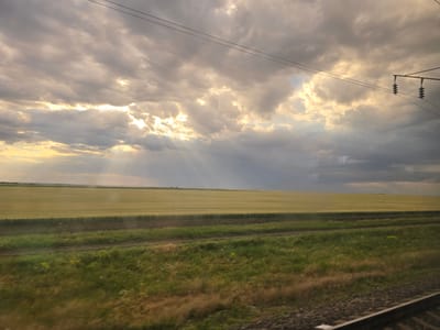 This image captures a wide-angle view of a rural landscape, likely from a moving vehicle like a train, looking out over agricultural fields under a dramatic sky. The scene is dominated by a vast, golden-green field of crops that stretches to the horizon. In the foreground, there is a strip of green vegetation with some dry grasses and wildflowers. The sky is filled with a mix of dark, brooding clouds and areas of bright sunlight breaking through, creating visible sunbeams. Two sets of parallel wires, presumably power lines, are visible stretching across the upper portion of the frame, with an electrical insulator hanging from one of them. The lower right corner of the image shows a portion of a train track, with ballast and rails visible. The overall impression is one of a dynamic weather system over an open, cultivated landscape. The location is identified as Peremozhne, Ukraine.