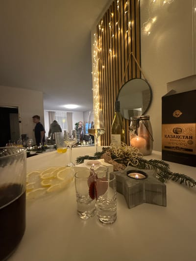 A table setting features glassware, including a large pitcher of dark liquid, wine glasses with lemon wedges, and small shot glasses. Decorative elements include a wooden box, a candle in a jar, and a star-shaped candle holder. Behind the table, a mirror hangs above a wall decorated with horizontal wooden planks and fairy lights. The reflection in the mirror shows a room with people gathered, illuminated by overhead lighting.