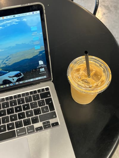A laptop with a Spanish keyboard is open on a black table. The laptop is displaying a screen with several folders, including "creando hábitos," "comiendo y corriendo," and "consultas." The laptop's power cord is plugged into the power outlet. A glass of iced coffee with a black straw sits next to the laptop. The location is Ciudad Santa Catarina, Mexico, at coordinates 25.658, -100.439. The scene is likely at a coffee shop or cafe.
