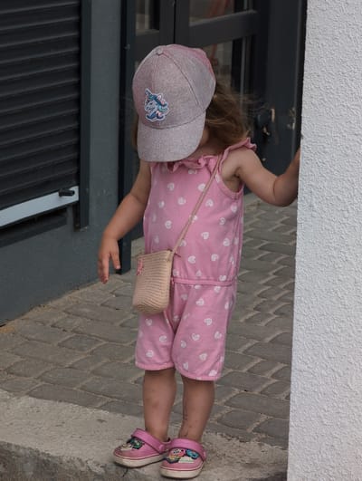 A young child, seen from the knees up, stands on a paved walkway in Perliavka, Ukraine. The child is wearing a pink jumpsuit with white hearts, a pink baseball cap with a unicorn emblem, and pink clogs. A small straw purse hangs from a string across the child's chest. The child's face is obscured by the cap, and their hair is visible peeking out from underneath. The child is positioned between a dark gray, slatted structure and a white textured wall.