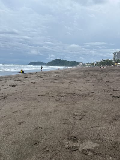 The image depicts a wide, sandy beach in Jacó, Costa Rica, under a cloudy sky. The dark sand is marked with numerous footprints. In the foreground, the sand is granular and slightly damp in places.
Further out, near the water's edge, several people are present. Two individuals, one in a yellow top and another in a darker garment, are seated in the shallow water, appearing to rest or converse. A lone figure, wearing a green shirt and dark shorts, stands on the sand near the water, looking towards the ocean. A small child is visible in the distance on the right, near the water.
The ocean exhibits moderate waves, with whitecaps breaking near the shore. In the background, a line of lush green hills rises, forming a natural backdrop to the beach. Beyond the hills, several multi-story buildings, likely hotels or condominiums, are visible along the coastline, indicating the presence of a developed resort area. White umbrellas and some beach chairs are scattered on the sand in front of these buildings. The overall atmosphere suggests a typical, overcast day at a popular beach destination.
