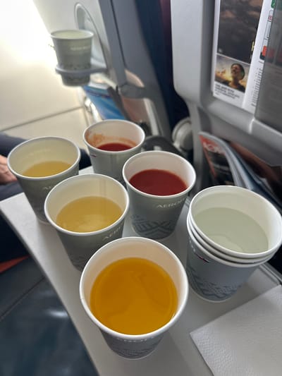This image shows a tray of drinks on an airplane. There are several paper cups filled with various beverages, including what appears to be orange juice, tomato juice, and water. Some of the cups have logos that suggest they are from Aeroflot. The tray is resting on a surface that is likely a fold-out table on the back of an airplane seat. In the background, another cup is attached to a seatback pocket. The lighting suggests it is daytime. The location specified in Maraldy, Kazakhstan is not depicted or inferable from the image.