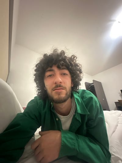 A man with curly dark hair and a beard lies on a white bed. He's wearing a green button-down shirt over a white t-shirt. A dark wood table is visible behind him. The room has white walls and what appears to be a white ceiling. He's looking directly at the camera with a slight smile. The photo is rotated slightly to the left.