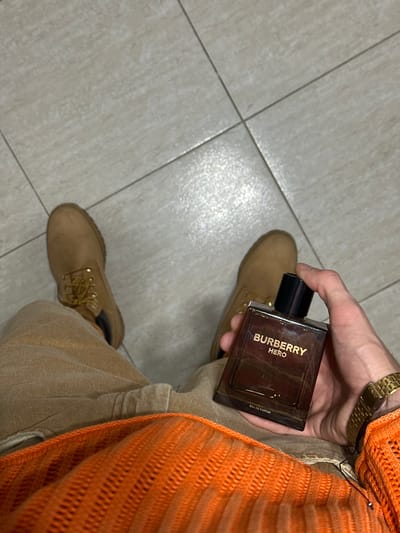 A person is holding a bottle of Burberry Hero Eau de Parfum in their right hand, with a gold wristwatch visible on the wrist. The person is wearing a beige outfit and tan-colored boots. Part of an orange knit sweater is visible in the upper right corner. The scene is situated on a light grey tiled floor. There is no indication of Los Palacios, Spain in the provided image.