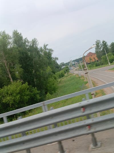 The image shows a rural road scene in Sibirsky, Russia, with a view from a highway guardrail. The guardrail, made of metal, is in the foreground, showing its layered structure and reflective markers. Beyond the guardrail, a paved road curves to the right, bordered by a grassy verge. In the distance, a two-story orange building with a red roof is visible, surrounded by lush green trees. Several concrete utility poles with streetlights are also present along the roadside. The sky is overcast with a pale grey hue, suggesting cloudy weather. The environment is predominantly natural, with a strong presence of trees and vegetation, indicating a location outside a dense urban area.