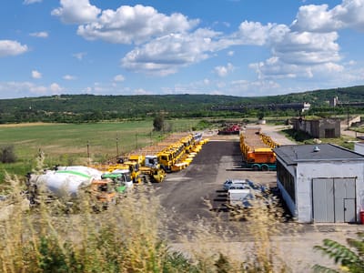 This is a daylight, wide-angle exterior shot of a construction equipment lot, possibly in the city of Yasen, Bulgaria. A large number of yellow cement trucks are parked in a long row on a paved area. Several other vehicles, including a white van, a silver car, and an orange cement truck, are also visible. In the background, there is a green, hilly landscape with trees and a blue sky with white clouds. A small building with a gray metal door and a white facade is in the foreground on the right side of the image. The foreground is blurred by tall, dry grass.