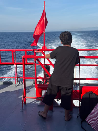 A young person stands on the deck of a boat, facing away from the camera and towards the open sea. They are wearing a black t-shirt and black shorts. A red flag is visible on a pole to the left of the person. The boat is moving, creating a wake in the water behind it. The sky is blue and clear, with distant land visible on the horizon. The deck of the boat is grey and made of metal plates. Red railings surround the deck. A black backpack and a checkered cloth are visible on the deck near the person's feet. The image is taken in the city of Dong Son, Vietnam, and the scene depicts a day at sea.
