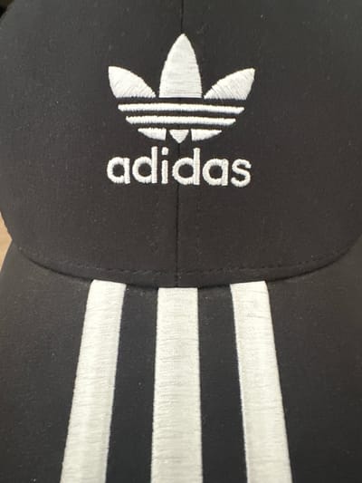 The image presents a close-up, angled view of a black fabric item, most likely a cap or hat, featuring white embroidered details. The main subjects are the embroidered logos and text on the black material.
On the left side of the frame, the iconic Adidas Trefoil logo is clearly visible in white embroidery, consisting of three parallel bars forming a stylized leaf. To the right of the logo, the brand name "adidas" is embroidered in white, lowercase sans-serif script. The texture of the embroidery threads is distinct, highlighting the craftsmanship of the stitching for both the logo and the text.
Extending across the right half of the image are three prominent, parallel white stripes, also embroidered onto the black fabric. These stripes are a characteristic design element associated with the Adidas brand. The black fabric of the item has a slight visible texture.
The lighting is soft and even, creating a clear contrast between the dark fabric and the bright white embroidery without harsh shadows. No people are visible, nor are there any discernible activities taking place. The background is out of focus, emphasizing the details of the clothing item. There are no specific location cues within the image itself to suggest Fulda, Germany. The overall impression is a detailed product shot focusing on branding elements.