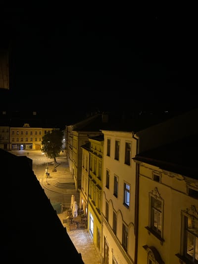 The image shows an outdoor nighttime view of a city street in Jihlava, Czechia. The scene is illuminated by streetlights and the lights from buildings. In the foreground, the dark silhouette of a rooftop juts into the frame from the left. Beyond this, a cobblestone street runs through the center of the image. On the left side of the street, a yellow building with multiple windows is visible, with one window glowing blue. Further down the street, a large tree stands in a small plaza. Several people are walking in the distance. On the right side of the street, a row of buildings with lit windows lines the pavement. In the middle ground, a cafe with outdoor seating is visible, with tables and chairs set up. The overall atmosphere is calm and quiet, suggesting a typical evening in a European city.
