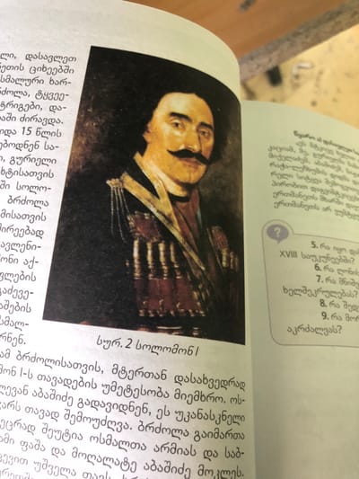 The image displays an open textbook, positioned diagonally, on a light-colored surface, likely a desk, in Ortabatumi, Georgia. The left page features a color portrait painting of a man with a prominent dark mustache and dark hair, wearing what appears to be traditional Chokha attire with cartridge belts across his chest. He looks slightly to the right, against a dark, indistinct background. Below the painting, a caption reads "სურ. 2 სოლომონ I" (Fig. 2 Solomon I) in Georgian script. The left page also contains multiple paragraphs of Georgian text. The right page features more Georgian text, including a section with a large question mark icon and a numbered list of questions, from 5 to 9, and the Roman numeral 'XVIII'.