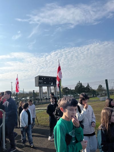 A daytime outdoor scene in ქალაქი ლანჩხუთი, Georgia, depicts a gathering of individuals, primarily adolescents and young adults, at what appears to be a sports facility. The sky is bright blue with scattered cirrocumulus clouds.
In the foreground, a young male wearing a green long-sleeved shirt with a white Nike logo is positioned slightly right of center, looking towards the right. His right hand is raised to his mouth, possibly chewing, while his left hand holds a dark object. To his immediate left and slightly behind, a young female in a white padded jacket and light-colored pants is visible. Other individuals in casual attire are scattered across the mid-ground, standing on a paved surface.
Two Georgian national flags, white with five red crosses, fly from flagpoles within the scene. One flag is positioned to the left of a prominent, dark-colored scoreboard structure, which stands on tall concrete supports in the mid-ground. The other flag is to the right of this scoreboard. A chain-link fence runs across the mid-ground, separating the standing crowd from a green, grassy field. In the background, beyond the fence and field, parked cars, trees, and distant hills or mountains are discernible.