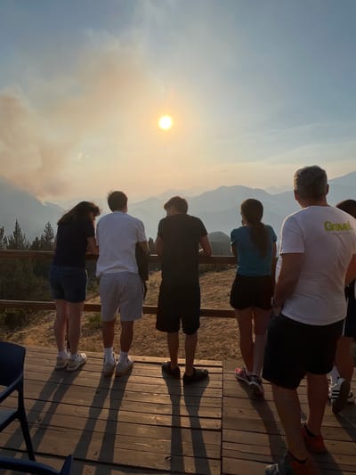 The image shows a group of six people standing on a wooden deck, looking out at a mountainous landscape. The sun is visible in the sky, partially obscured by smoke, suggesting a possible wildfire in the distance. The people are casually dressed in t-shirts and shorts. The person on the far right is wearing a white t-shirt with green lettering that reads "Gravel". The location is identified as San Juan de Plan/San Chuan de Plan, Spain. The overall lighting suggests it is late afternoon or early evening, with long shadows cast across the wooden deck. The environment appears to be a natural, possibly elevated, viewpoint overlooking valleys and mountain ranges.