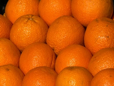 A close-up, eye-level, medium shot captures a pile of bright orange oranges, some appearing slightly bruised, filling the frame. The lighting is bright, highlighting the textured skin of the fruit. The background is out of focus, with no discernible details about the surroundings or any indication of Shalikiantkari, Georgia. There are no people or actions depicted in the image.