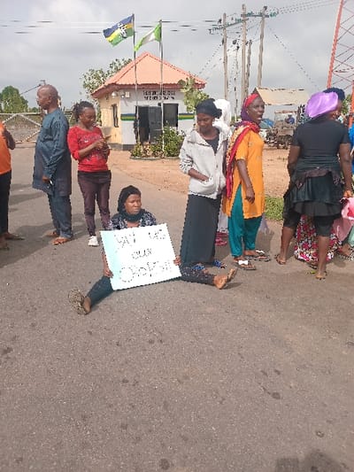 The image shows a group of people in front of a building with a sign that reads "Federal Correctional Service." There are several people standing and one woman sitting in the middle of the road, holding a sign that reads "We want our Boy Back." The woman is wearing a black hijab and black clothing. Behind her are other women wearing hijabs and brightly coloured clothing. There is a man wearing a white shirt and a woman wearing a green headscarf standing behind the seated woman. There are power lines in the background and a green bush on the left. The scene appears to be in front of a correctional facility in Ungwan Tanko, Nigeria.