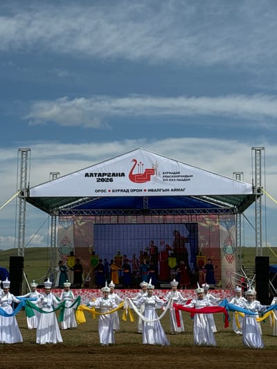 An outdoor stage setup is visible, featuring a white canopy with Cyrillic text and a red graphic of a bird. The text translates to "ALTARGAHANA 2026" and indicates the event is in Orongoi, Buryatia, Russia, specifically for the XVI Naadan. A group of individuals, appearing to be performers dressed in traditional white costumes and hats with colorful sashes, are positioned in the foreground, some holding and extending long, flowing sashes. Behind them, on the stage, other performers are visible, some standing and others engaged in what appears to be a traditional dance or performance, set against a backdrop displaying various images and patterns. The stage is supported by metal scaffolding and is set on a grassy, uneven terrain under a partly cloudy blue sky.