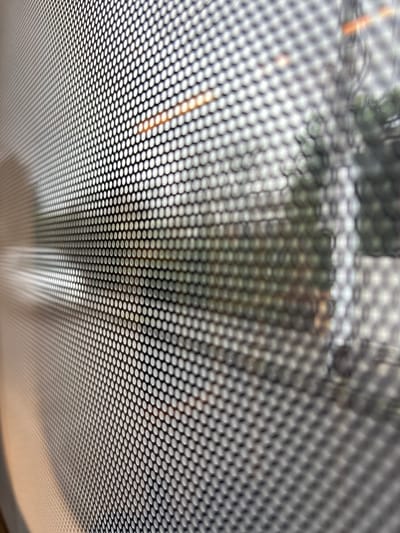 This is a close-up, abstract photograph of a perforated screen or material, viewed from an oblique angle. The screen is composed of a dense pattern of small, circular perforations that create a honeycomb-like effect. The perforations are more in focus in the foreground and become progressively blurred towards the background. Through the screen, a blurry, out-of-focus scene is visible, suggesting it is a window or barrier. Hints of greenery, possibly trees or bushes, can be discerned, along with some warm-toned light sources, possibly streetlights or reflections. The image does not depict any specific identifiable landmarks, people, or activities that would allow for a definitive location within Brunswick East, Australia. The overall impression is one of texture and obscured vision.
