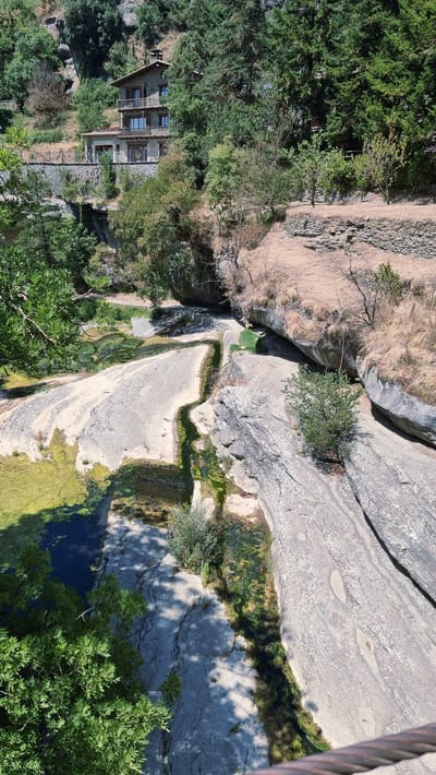 The image shows a scenic view of a natural landscape near Rupit i Pruit, Spain. A multi-story stone building with wooden balconies is situated on a hillside, surrounded by lush green trees and vegetation. Below the building, a shallow river flows over smooth, light-colored rocks, with patches of green algae or moss visible in the water. The terrain is rugged, with rocky outcrops and dry grasses on the slopes. The scene is bathed in bright sunlight, casting shadows on the ground and rocks.