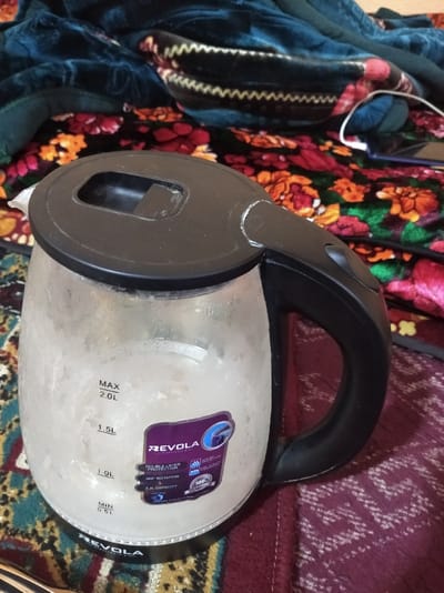 A clear glass electric kettle, branded "REVQLA" with a black lid and handle, is centrally positioned on a patterned textile surface. The kettle contains a visible amount of liquid and exhibits internal condensation, with volume markings ranging from 0.5L to a 2.0L MAX line. A purple manufacturer's label on the kettle details features such as "DOUBLE-LAYER PROTECTION," "360° ROTATION," and "2.3L CAPACITY." The textile directly beneath the kettle features a geometric pattern in shades of maroon, beige, and dark green. Behind the kettle, the surface transitions to a floral-patterned textile in red, orange, and green on a dark background. Further back, a dark blue or green textured fabric with a distinct light-colored zig-zag pattern is visible. A white charging cable and a dark-colored smartphone are partially visible in the background, resting on the floral-patterned textile. No individuals are present, and no actions are being performed. The scene is an interior setting in Asaka, Uzbekistan.
