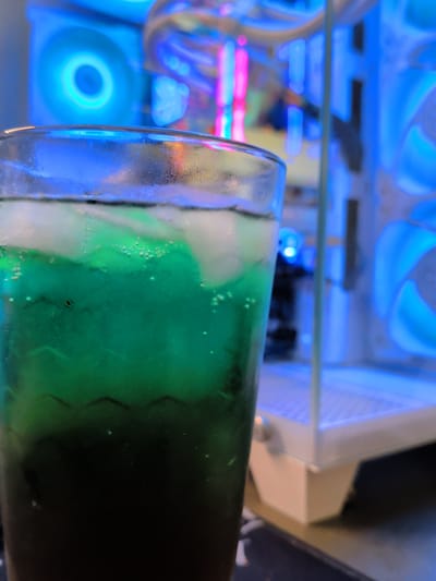 A close-up, low-angle shot captures a glass of green liquid with ice cubes and condensation, placed in front of a brightly lit computer case. The computer case emits a cool blue and pink glow from its internal lighting and fans. The background is blurred, emphasizing the drink in the foreground. The scene is indoors, and there is no indication of the city of Galiche, Bulgaria.