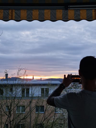 A person stands on a balcony in Rheinfelden, Switzerland, taking a picture with their smartphone of a sunset viewed from the balcony. The sunset casts an orange glow across the sky and on the buildings below. The person is silhouetted, revealing dark hair, a light-colored t-shirt, and a watch on their left wrist. Their left arm shows a tattoo. The view includes a building with white-framed windows and a light orange facade. A bare tree with budding leaves is partially obscuring the view of the building. The balcony is sheltered by a yellow and gray striped awning. The sky shows light gray and purple hues. The overall scene suggests a residential setting in Rheinfelden during the transition between day and night.