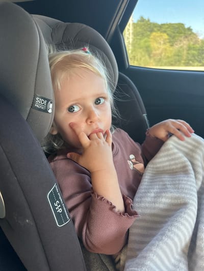 A toddler is lying in a gray car seat, partially covered by a striped gray and white blanket. She wears a mauve long-sleeved shirt with a cartoon design. Her blonde hair is pulled back with colorful hair ties. Her eyes are wide, and she has a finger in her mouth. The car seat is labeled "SIDE PROTECT" and "S.I.P." Outside the car window, green trees and a bridge-like structure are visible. The location is unknown.