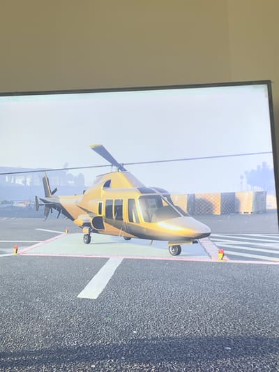 This image depicts a digitally rendered, metallic gold helicopter with black accents, parked on a helipad marked with red and white lines. The helicopter has two visible landing gear wheels on the left side and one on the right. In the background, there are stacks of pallets wrapped in plastic, and beyond that, a blurry treeline and a light-colored building. The scene is set on an asphalt surface. The image does not depict the city of Vejprty, Czechia.