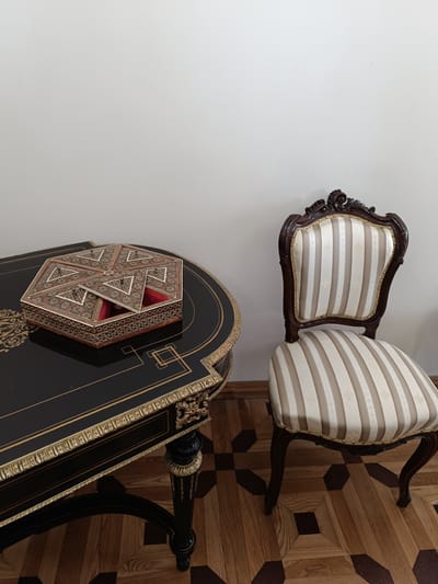 The image depicts an indoor scene featuring a table and a chair. The table is dark, ornate, and has gold detailing. On the table sits a decorative octagonal box with intricate patterns and a lid that is slightly ajar. To the right of the table is a wooden chair with a carved dark wooden frame and a cushioned seat and backrest upholstered in a striped fabric of beige and white. The floor is a patterned parquet with alternating dark and light wood planks. The background is a plain, light-colored wall. The scene appears to be set in a room with a classic or antique aesthetic. There are no people visible in the image.
