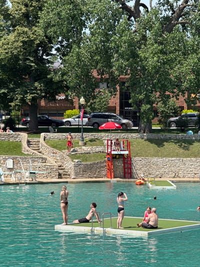 The image depicts a crowded outdoor swimming pool on a sunny day, likely in Naperville, Illinois, given the presence of a "445 W. JACKSON" sign on a brick building in the background, a common street name in the city. The pool has vibrant turquoise water and is surrounded by stone walls and grassy slopes. Several people are visible in and around the water. In the foreground, a floating platform with a green turf surface hosts a woman in a bikini and a man in swim trunks sitting together. To the left of the platform, another woman in a bikini stands, and a man is seated in the water. Further back, a person is swimming. On the far right of the pool, another floating platform has a woman in a bikini and a man sitting together. In the background, a lifeguard stand with a red ladder and umbrella is positioned above a stone staircase. A man in red swim trunks and a blue cap stands at the top of the stairs, holding a rescue tube. Behind the pool, a brick building with large windows is visible, along with trees and parked cars. A street light and some signage are also present. The scene suggests a recreational activity, with people enjoying the warm weather at a public pool.