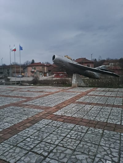 The image depicts an outdoor scene in the city of Kamenets, Bulgaria, under a uniformly overcast, grey sky. In the midground, a metallic grey jet aircraft, resembling a Mikoyan-Gurevich MiG-15 due to its nose intake and swept wings, is mounted on a light-colored, angular concrete pedestal. The aircraft is angled upwards and to the right, simulating a takeoff or climbing posture. To the left of the aircraft, three flagpoles are visible; the left two display the Bulgarian national flag (white, green, red horizontal stripes), and the rightmost displays a solid blue flag. Behind the flagpoles and aircraft, a residential area features several two-story buildings with light-colored facades and reddish-brown tiled roofs, interspersed with bare trees and utility poles. A silver sedan is partially visible, parked on a paved surface among these structures. The foreground consists of a plaza paved with a repeating pattern of light grey square tiles, each containing an irregular mosaic of stone fragments, bordered by darker, reddish-brown rectangular paving stones. No individuals or actions are present in the scene.