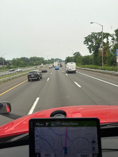 The image shows a view from the driver's perspective of a semi-truck on a highway. The truck has a red hood and a black side mirror visible. A GPS device is mounted on the dashboard, displaying a map with the destination "White Plains". The highway is multi-lane, with other vehicles including a black SUV, a white van, and a blue car. Road signs are visible, including one for Interstate 87 South. The environment appears to be a suburban or rural area with trees lining the highway and an overcast sky. Based on the GPS display showing "White Plains" and the Interstate 87 South sign, this scene is likely in the vicinity of Danbury, Connecticut, USA, as this highway passes through or near that city.