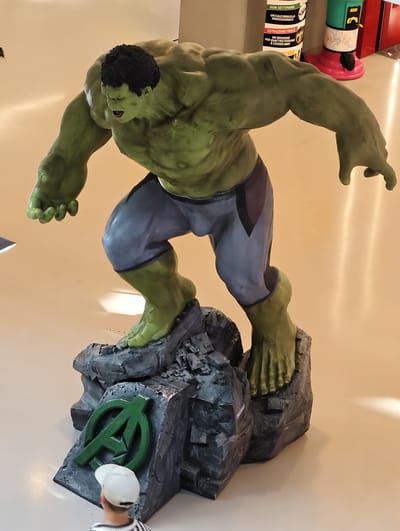 An imposing, life-sized statue of the Hulk, a green-skinned, muscular superhero, stands on a rocky, grey base. The Hulk is depicted in a dynamic pose, leaning forward with one arm extended and his mouth open in a roar. He wears grey shorts with darker accents. The base of the statue features the green Avengers logo. In the foreground, partially obscured by the base, is a person wearing a white baseball cap and a black and white striped shirt, looking up at the statue. The background appears to be an indoor public space with a polished floor and a hint of some commercial displays or machinery on the right. This scene is located in Nola, Italy.