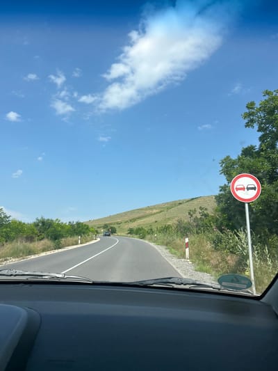 This image presents a first-person perspective from within a vehicle, looking forward onto a winding asphalt road under a bright, clear blue sky with scattered white cumulus clouds. The scene is situated in the area of Isperihovo, Bulgaria, in a rural or semi-rural environment characterized by rolling hills covered in green and dry vegetation, suggesting a summer or late spring season.

The dark grey asphalt road curves to the left in the distance, marked by a solid white center line. On the right side of the road, a white and red reflective delineator post is visible, with gravel and natural vegetation like tall grasses and flowering thistles growing on the shoulder. Further down the road, a dark-colored sedan is visible, also traveling in the same direction.

Prominently displayed on the right side of the road, a circular regulatory road sign is affixed to a metal pole. The sign features a red border on a white background, depicting two cars side-by-side: a red car on the left and a black car on the right. This sign typically indicates a "No Overtaking" or "No Passing" zone.

In the immediate foreground, the dark dashboard and a windshield wiper blade are visible, framing the view through the vehicle's windshield. A green oval sticker is adhered to the lower right portion of the windshield. No individuals are explicitly visible in the scene.