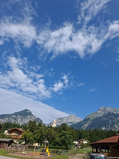 This image depicts a scenic view of the Austrian Alps from the city of Münster. In the foreground, a children's playground with a yellow slide and wooden fencing is visible. Beyond the playground, several houses with traditional Austrian architecture are nestled among lush green trees. The backdrop is dominated by the majestic, rugged peaks of the Alps, with some parts showing exposed rock and others covered in dense coniferous forests. The sky above is a vibrant blue, dotted with scattered white clouds. The overall scene suggests a peaceful, rural setting with a focus on outdoor recreation and natural beauty.