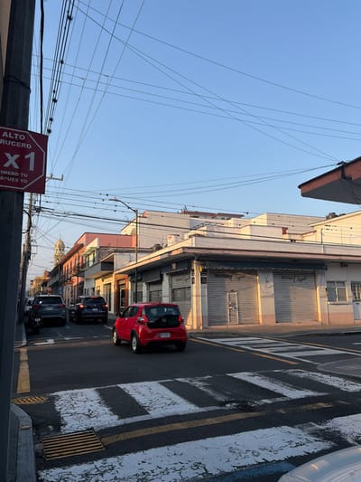 A street scene captures buildings with closed metal shutters, and a red car parked beside a crosswalk. Several cars follow down the road. Power lines crisscross the sky above. A stop sign reads "Alto Crucero X1" with further Spanish text. The perspective is skewed, as if viewed at an angle, and the sky is a clear blue.