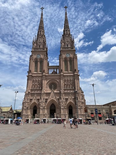A tall, ornate cathedral with two prominent spires dominates the scene. It's likely built from light brown stone. Two clocks are visible on the towers. The sky is blue with wispy clouds. People walk along a paved area adjacent to the building. There are a couple of lampposts in view. Part of a building can be seen to the right of the cathedral and it appears to be commercial or mixed use, with shops and stalls set up in front.
