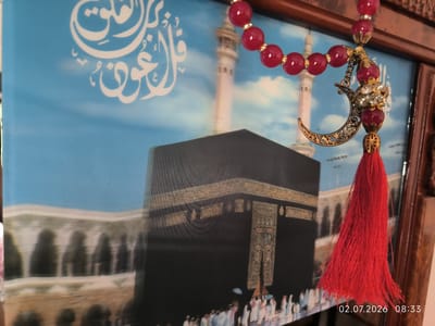 A framed image depicts the Kaaba in Mecca, Saudi Arabia, with Islamic calligraphy in white on a blue sky background. A decorative string of red beads, a crescent moon charm, and a red tassel hangs in front of the image, partially obscuring it. The frame is made of dark wood. A timestamp "02.07.2026 08:33" is visible in the lower right corner. There are no people or animals present in the image itself, and no specific location in Kattasu, Uzbekistan is indicated.