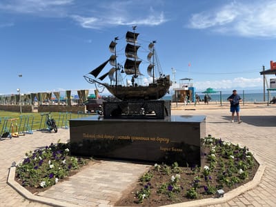 This is a medium, eye-level, outdoor shot featuring a sculpture of a ship. The sculpture is made of dark metal and is mounted on a dark, polished stone pedestal. The pedestal has an inscription in gold lettering. The ship sculpture has multiple masts and sails, and intricate details representing ropes and rigging. It is positioned in a landscaped area with a flowerbed of purple and white flowers around the base of the pedestal.

The scene appears to be a public space, possibly a waterfront promenade or park, given the presence of a beach, tents with drapes, and people in the background. To the right of the sculpture, a man is walking away from the camera, wearing blue shorts and a dark shirt. He is carrying a bag. In the background, there are beach umbrellas and tents, a lifeguard tower, and mountains in the far distance. The sky is blue with some scattered white clouds, suggesting it is daytime and fair weather.

The inscription on the pedestal reads: "Поймай свой ветер - оставь сомнения на берегу..." followed by "Генрих Балян". This translates to "Catch your wind - leave your doubts on the shore..." and the name "Heinrich Balian". The overall mood is peaceful and scenic, with a touch of artistic inspiration from the ship sculpture and its accompanying quote. The presence of recreational beach facilities and people suggests a leisurely atmosphere.