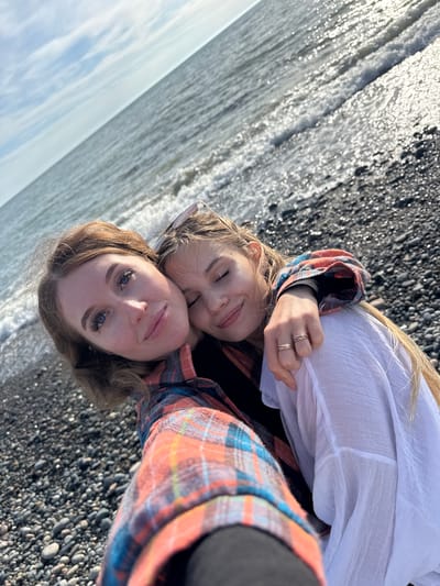 Two Caucasian women pose close together on a black pebble beach, likely taking a selfie. The woman in the foreground has light brown hair and blue eyes. The other woman, partially obscured behind her, has long blonde hair and is wearing sunglasses pushed up on her head. The background shows the ocean with shimmering sunlight reflecting off the water. The location is probably a beach near the city of Gulrypsh, Abkhazia, as indicated in the prompt.
