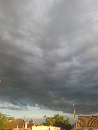 The image shows a dramatic sky with dark, heavy clouds on the left and lighter, scattered clouds on the right. A faint rainbow is visible against the darker clouds. Power lines crisscross the sky. In the foreground on the right, parts of buildings are visible, including a wooden-sided structure with a corrugated roof, and another building with a red roof and lighter siding. Trees with green foliage are also present, suggesting a rural or suburban setting. The light suggests it is either early morning or late afternoon, with the sun trying to break through the storm clouds. The overall mood is one of impending weather, perhaps a storm passing or approaching. There is no visible text in the image. The scene appears to be outdoors, possibly in Atbasar, Kazakhstan, given the prompt's location context.